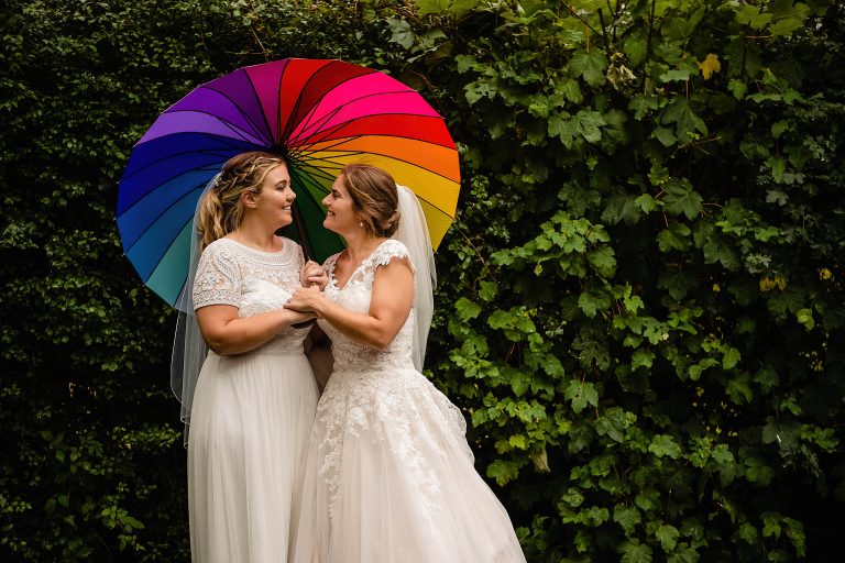 two brides standing under a rainbow umbrella. village hall wedding photography by emma and rich. LGBTQ+ wedding photographers.