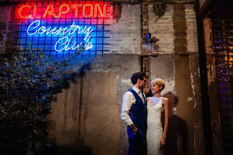 a bride and groom pose beneath a neon sign of the clapton country club wedding photography by emma and rich