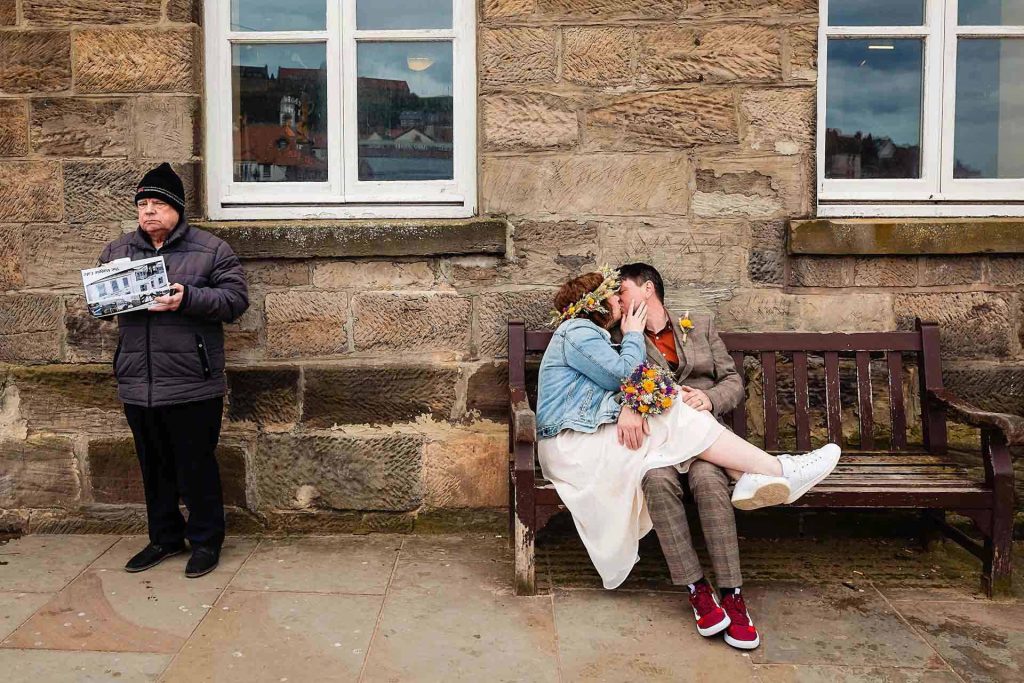 a newly married couple kissing on a bench next to a man eating fish and chips. Whitby elopement wedding photography by emma and rich